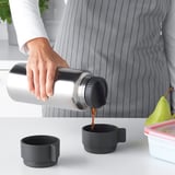 Persona abocant cafè d'un termo metàl·lic a una tassa negra sobre una taula blanca, amb una altra tassa i un recipient de menjar a prop.