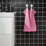 La tovallola VÅGSJÖN de color rosa brillant penja a la paret, rajoles fosques contrastades, dos ganxos blancs. Cotó suau, mida mitjana, plegat, bany modern.