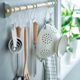 Els estris de cuina penjaven en ganxos. Es veu un zèster blanc amb mànec de fusta, tasses de mesura, espàtula de fusta i colador blanc.