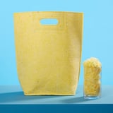 La bossa groga gran de VÄXELBRUK, feta de fibres reciclades, s'alça al costat d'un got més petit de trossos grocs sobre una superfície blava.