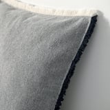 Primer pla de la funda de coixí teixida amb vores de serrells. Teixit gris clar amb lleugera textura. Els serrells són de color blau fosc i blanc.