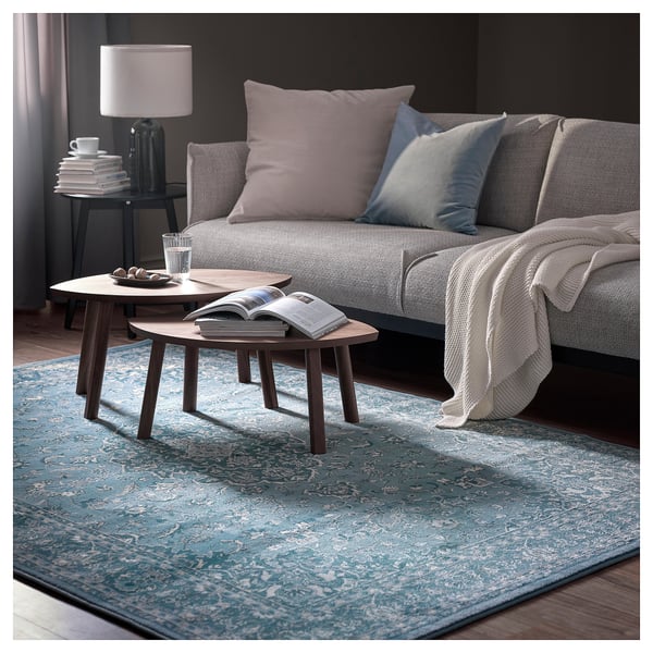 Sala d'estar moderna amb sofà gris, coixins blaus, taules de centre de fusta i catifa blava estampada.