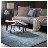 Sala d'estar moderna amb sofà gris, coixins blaus, taules de centre de fusta i catifa blava estampada.
