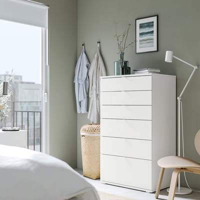Dormitori modern amb cofre blanc VIHALS, parets de color verd clar, cadira de fusta, llum, tèxtils i estampat natural.