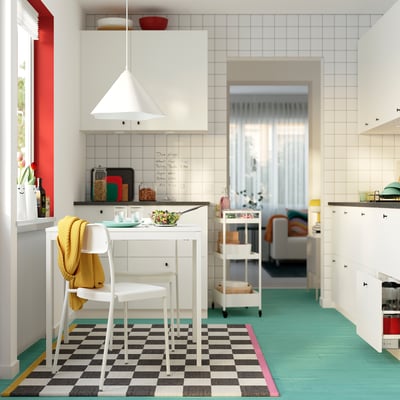 Taula de menjador blanca i minimalista amb cadires, situada en una cuina de colors. Manta groga brillant, accents vermells.