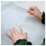 Person hand-embroidering beige cushion cover with intricate white patterns. Green yarn and fingers visible, focusing on detailed craftsmanship.