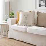 White ÅKERNEJLIKA embroidered cotton cushion with blue flowers on sofa.