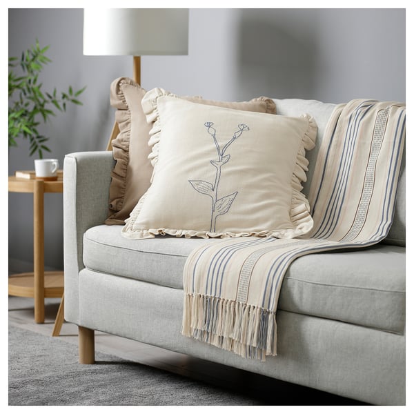A cozy living room with a sofa, ÅKERNEJLIKA cushion, striped throw, lamp, and plant.