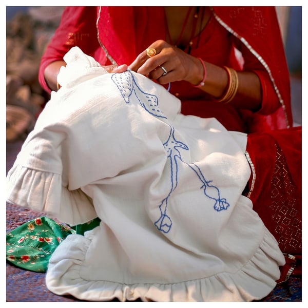 Woman in red dress hand-embroidering white, ruffled cushion cover with blue patterns.
