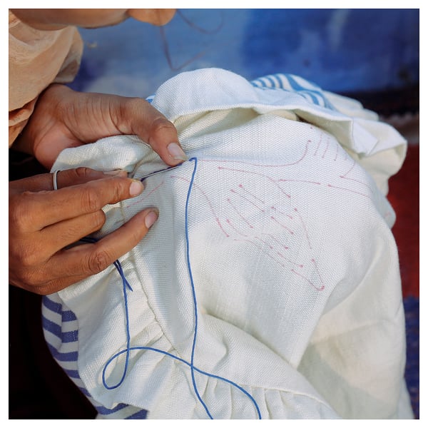 Person hand sewing white IKEA ÅKERNEJLIKA cushion cover with blue thread.