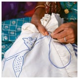 A person hand-sews a white, blue-embroidered cushion with intricate patterns.