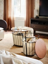 Two rustic storage tables with beige fabric, wooden tops, and black metal frames hold folded blankets near a window.