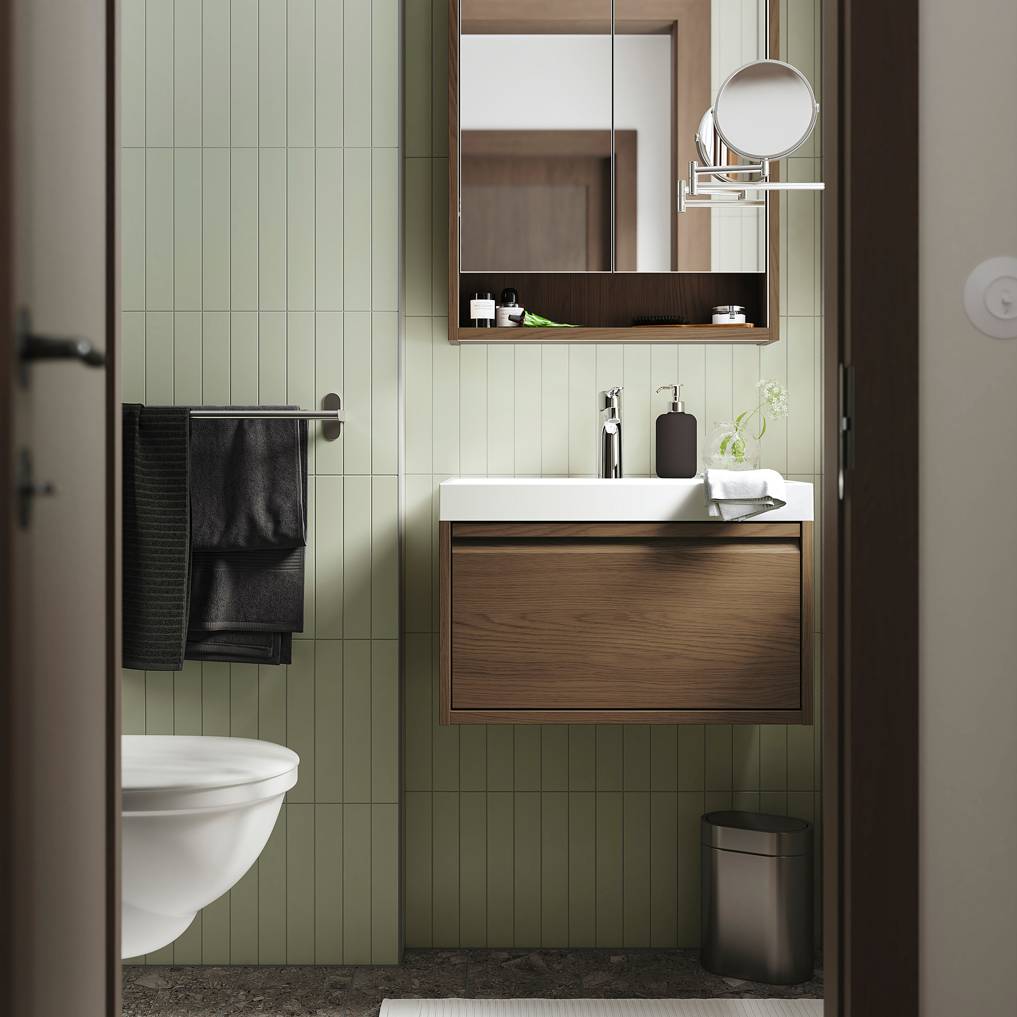 Modern bathroom with light green tiles, white sink, dark wood cabinet, black towels, and metal accents. Vanity fits neatly beneath a large mirror.