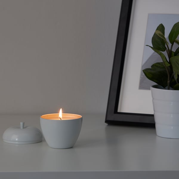 A small lit candle, ANSPRÅKSLÖS in light blue, sits on a surface beside its lid. Next to a small plant and a framed picture.