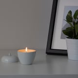 A small lit candle, ANSPRÅKSLÖS in light blue, sits on a surface beside its lid. Next to a small plant and a framed picture.