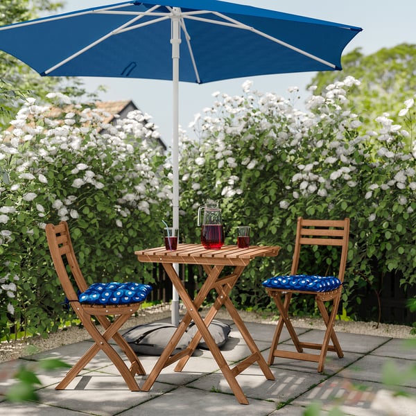 Small outdoor seating set with blue umbrella, table, and chairs near blooming bushes.