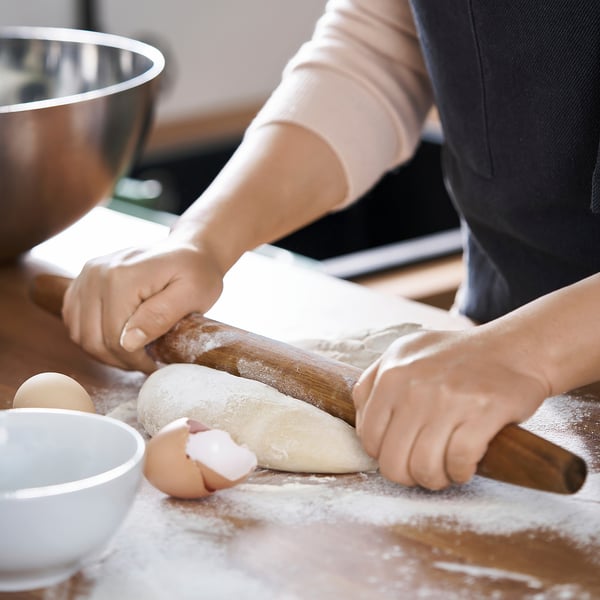 Two people are rolling out dough using a wooden handle-free rolling pin on a floured surface. A child cracks an egg into a bowl nearby.