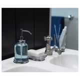 Chrome soap dispenser on white sink, with glass & metallic pump, plus soap dish & toothbrush holder.
