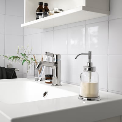 Modern chrome bathroom sink soap dispenser sits right beside faucet, smaller than the glass vase with green plant.