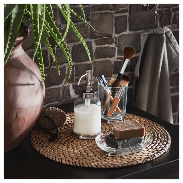 Bathroom counter with glass soap dispenser, small plants, makeup brushes, and a soap dish. Stone wall background.