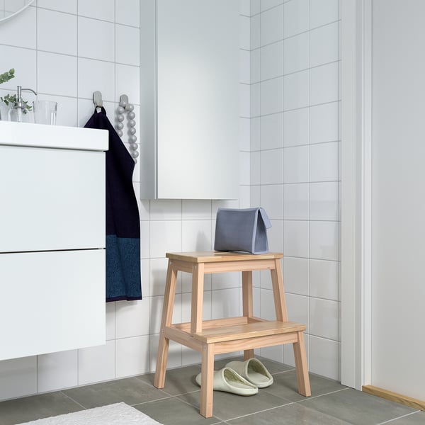 Minimalist bathroom with white tiled walls. Wooden BEKVÄM step stool holds blue towel & slippers.