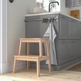 Wooden BEKVÄM step stool beside gray kitchen cabinets. Small, light-colored, stable.
