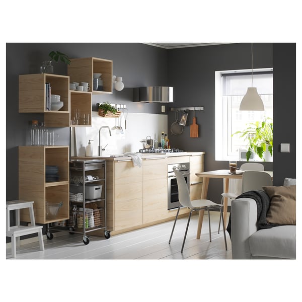 Modern kitchen with wood cabinets, white appliances, and gray sofa. Open shelves display dishes. Step stool, wooden chairs, window, and plant.