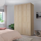 Bergen sliding wardrobe in light oak. Tall, narrow, modern panels. Bed, plants, rug nearby.