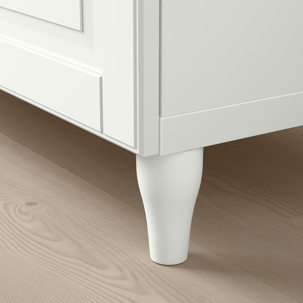 Modern white cabinet with sleek, tapered legs on light wooden floor.