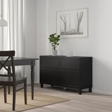 Minimalist dining area with black BESTÅ storage unit, wooden table, chair, and potted plant.
