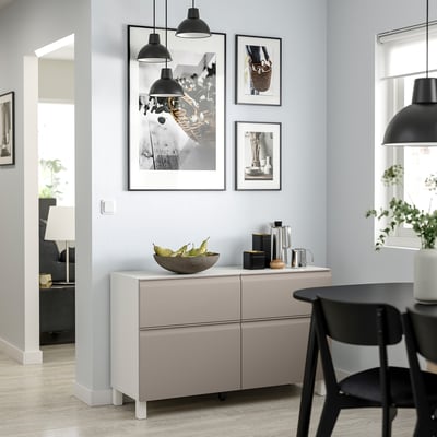 Modern dining area with light gray BESTÅ sideboard, black chairs, and pendant lamps. Artwork and fruit bowl on the sideboard.