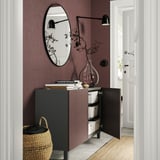 Dark brown BESTÅ cabinet, with open doors showing white organizers, against purple wall. Topped with books, a vase, and a large round mirror.