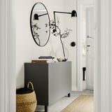 Modern entryway with BESTÅ grey storage unit. Large round mirror, wall lamps, and decorative vase with branches.