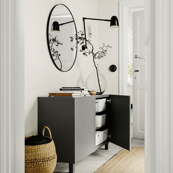 Modern entrance with black BESTÅ storage unit, white walls, round mirror, black wall lamps, woven basket, and glass vase with branches.