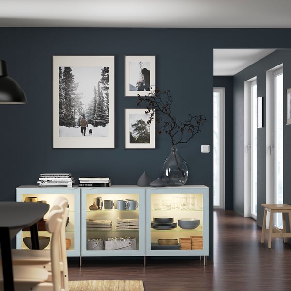 A modern dining room a light blue BESTÅ sideboard against grey walls. The sideboard holds dishes and decor. Black and white photos hang above it.