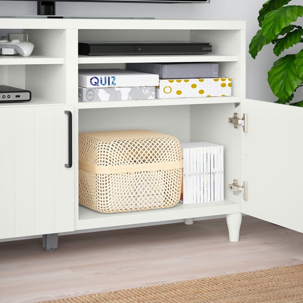 White BESTÅ tv bench with adjustable shelves, storing wicker basket, game boxes, and books. Soft-closing hinges visible.