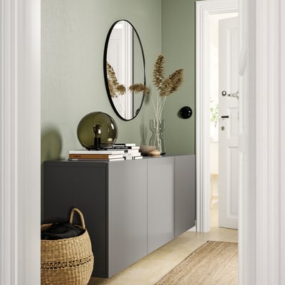 Modern entryway with light green walls, white door, gray BESTÅ cabinet, round mirror, woven basket, lamp, books, and dried flowers.