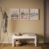 Three city prints: new york, london, paris on beige wall; white bench with hat and scarf; coat rack with jackets.