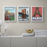 Three posters of rome, brooklyn bridge, and paris hang above a grey sideboard. The sideboard holds a white lamp, a gold bowl, and stacks of books.