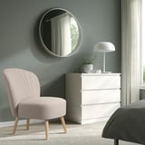 A stylish, light pink BILLHAMN armchair next to a white chest of drawers. The chairs compact, curved design complements the minimalist bedroom decor.