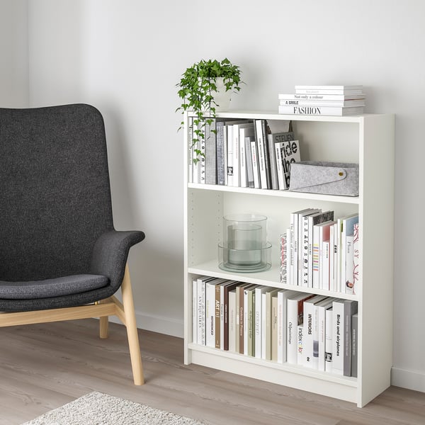 White BILLY bookshelf with 3 shelves, holding books, candle, and plant. Gray chair beside it, fits neatly in corner.