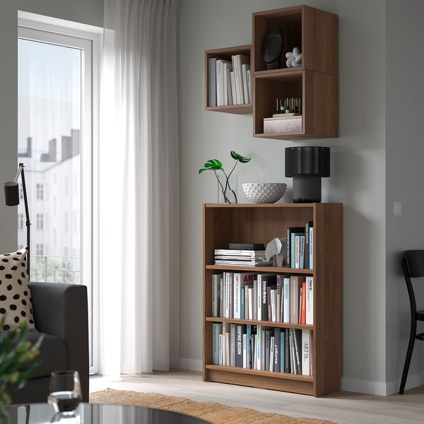 Wooden bookshelf with books, lamp, plant, beside window. Gray wall, two upper shelves.