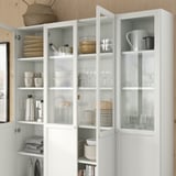 A white BILLY bookcase with glass doors, displaying neatly organized dishes, glassware, and kitchen items on shelves.