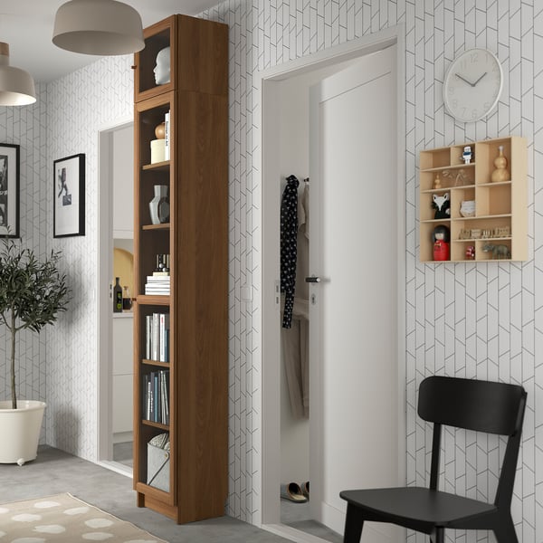 Modern entryway with tall wooden BILLY bookshelf, black chair, geometric walls, white door, clock, and potted plant.