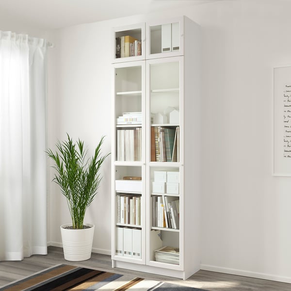 A tall white BILLY bookcase with glass doors, holding books and decor, against a white wall. A large plant sits beside it.