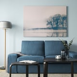 Large BJÖRKSTA frame picture, calm nature scene with trees, 32x45 inches, blue tones. Hangs above blue sofa, with coffee setup nearby.
