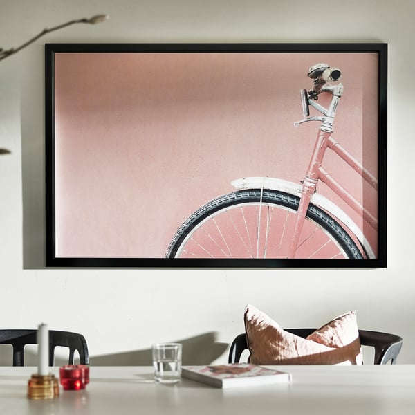 Pink bicycle with white seat, soft pink background, table with glass, candle holders, book, cushions.