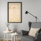Vitruvian man in BJÖRKSTA frame, gray wall, wingback chair, desk lamp, small tables with cup and dried flowers.