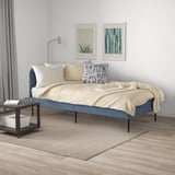 Blue daybed with beige bedding against white wall, flanked by decor, under grey curtains. Cozy, compact.