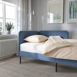 Blue upholstered bed frame with headboard and white pillows, positioned near a window with a plant on the sill and art on the wall.
